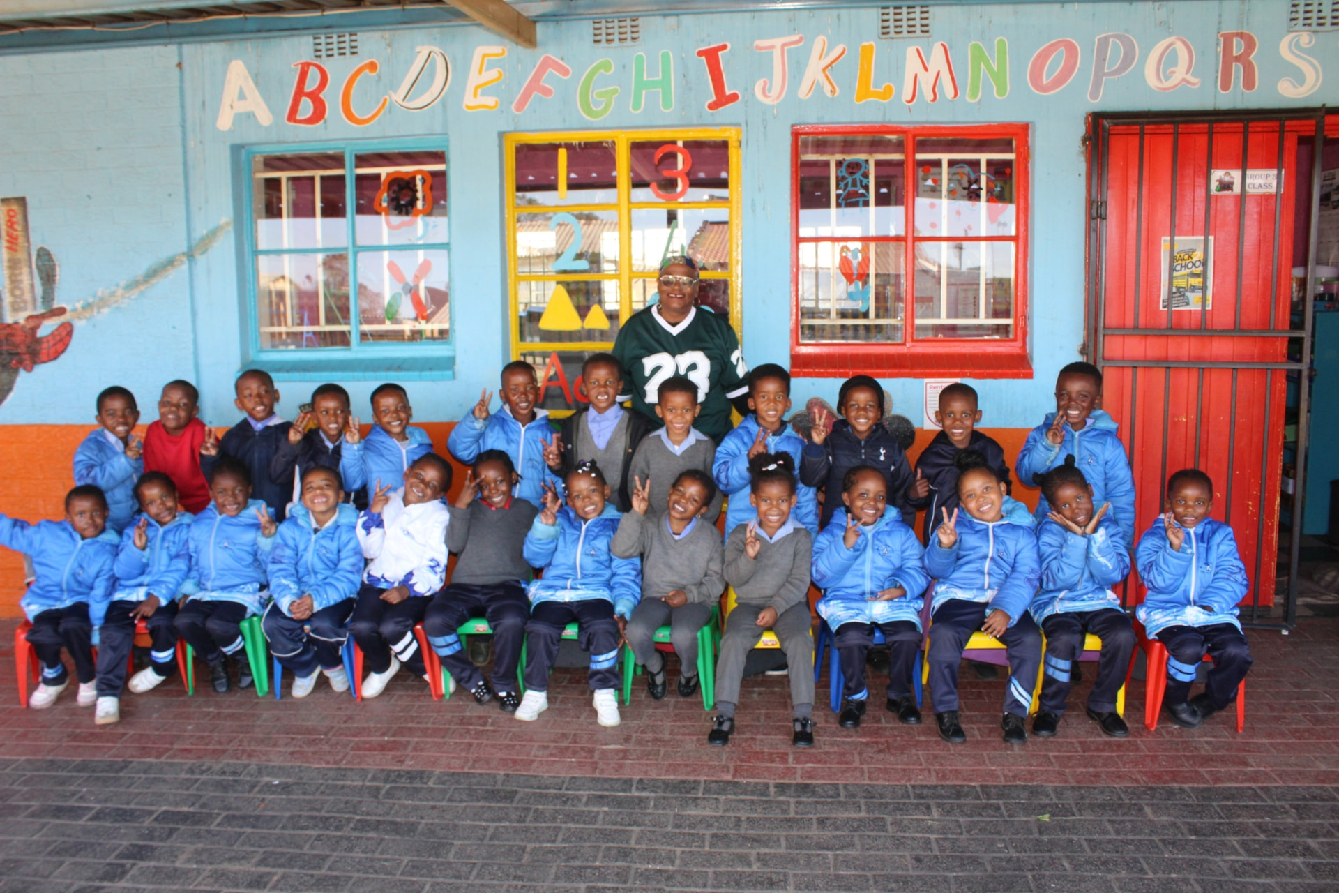 Matla Pre School Grade R class in blue jackets seated with their teacher in front of the painted alphabet wall
