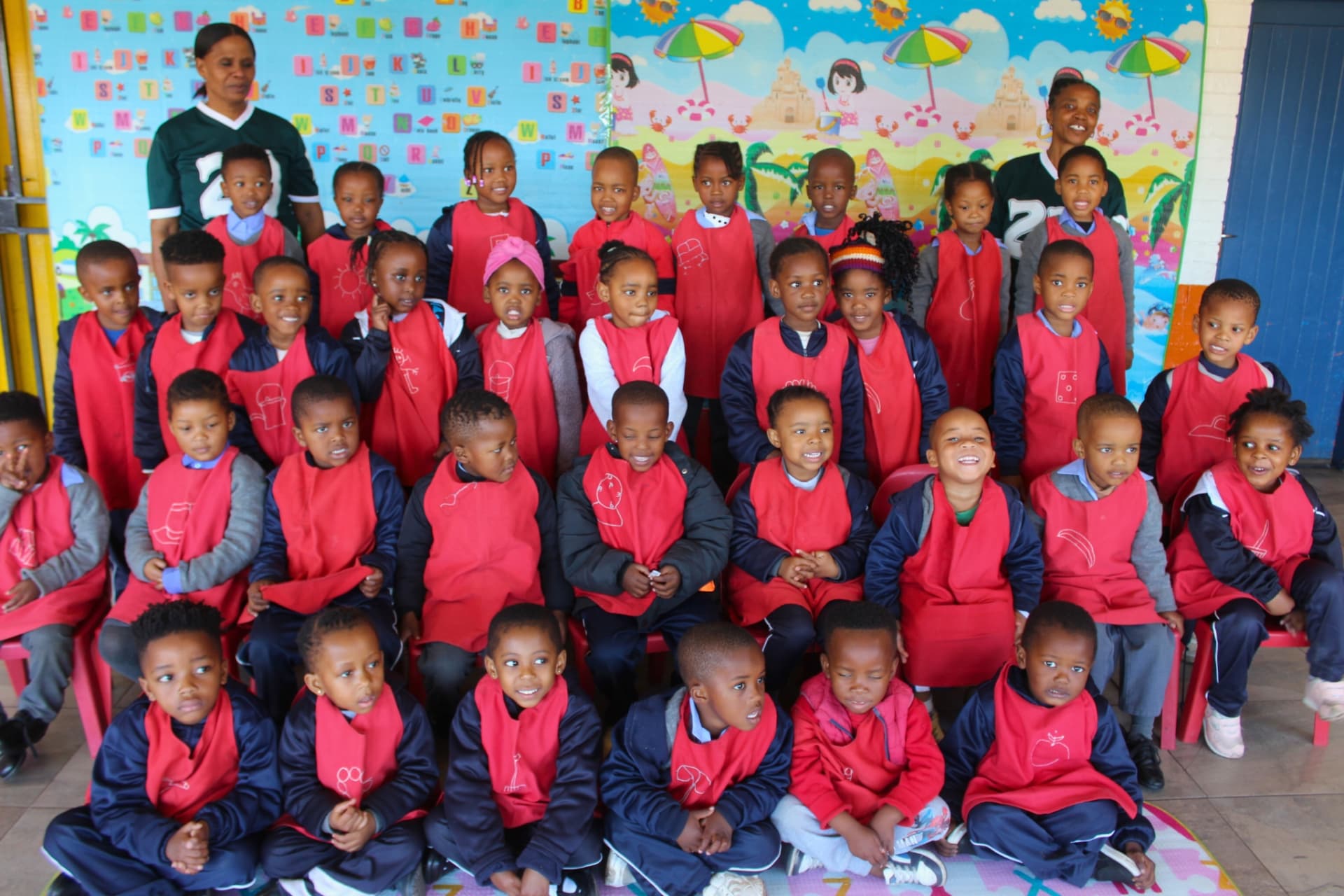 The red group class photo with their teachers in front of the alphabet backdrop