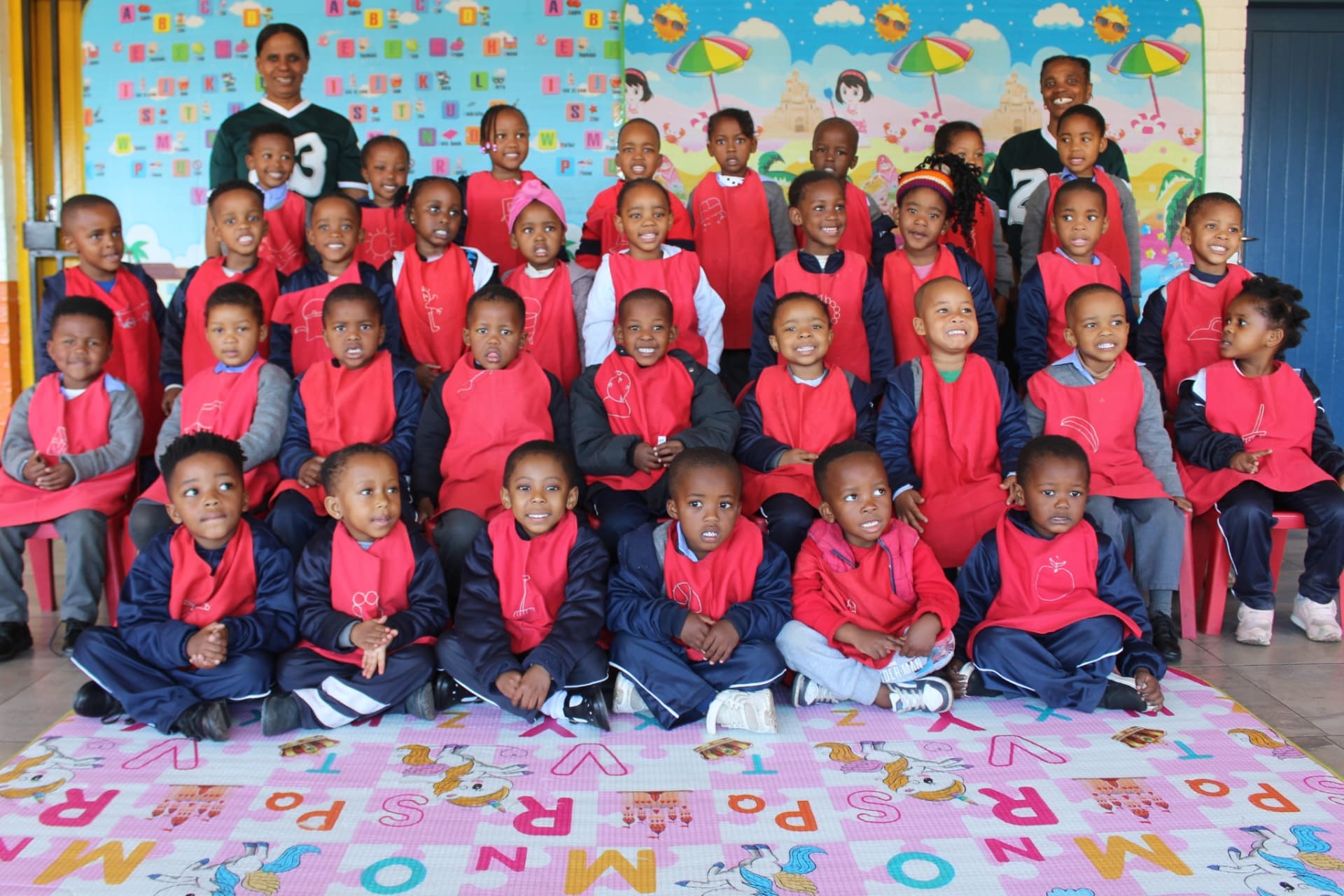 Matla Pre School Pre-Grade R class in red aprons seated on the alphabet mat with their teachers